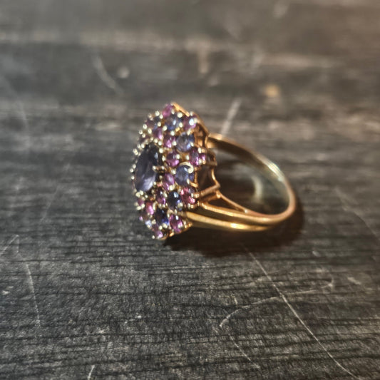 A gold ring with a cluster of pinkish-purple stones and a central oval purple gemstone, resting on a dark textured wooden surface.
